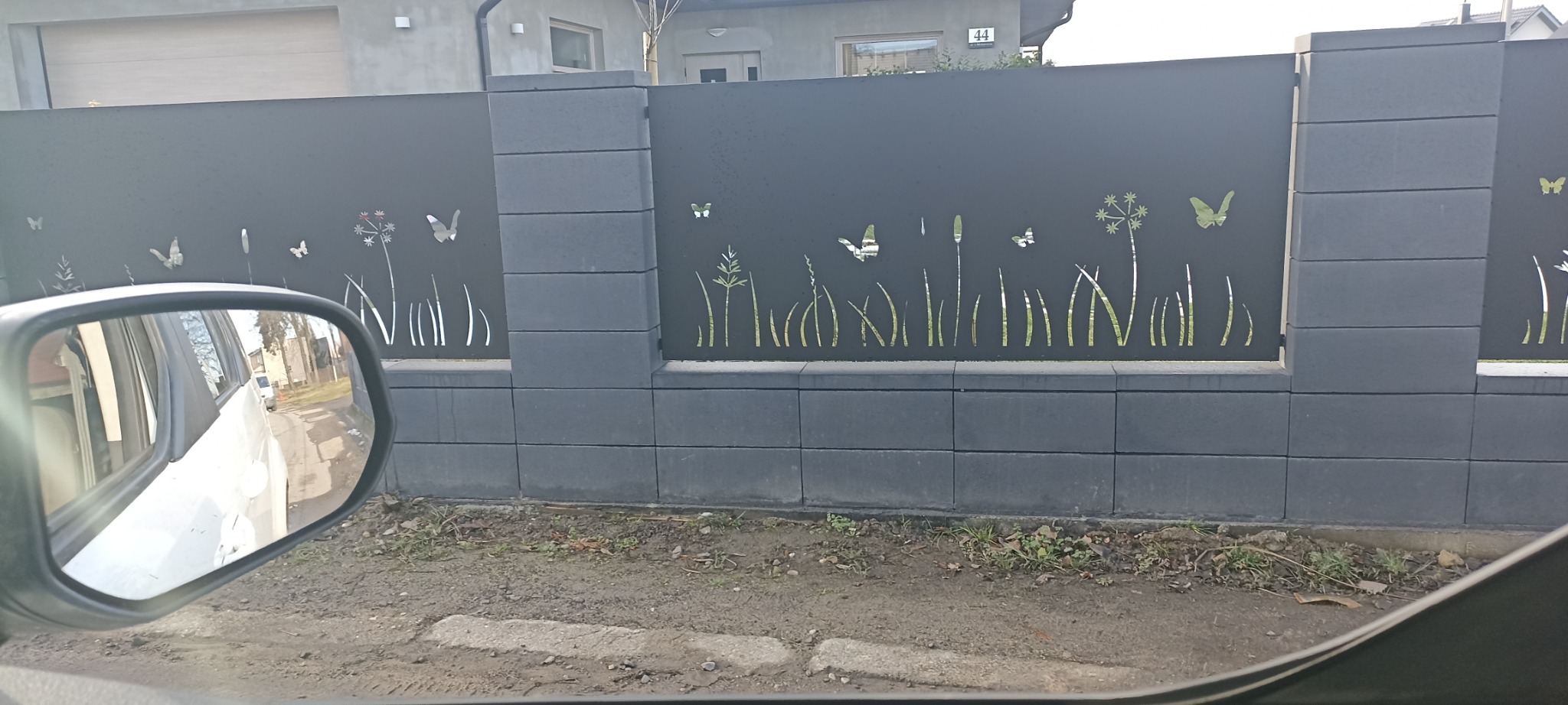 Nowoczesne ogrodzenie panelowe w kolorze grafitowym z wycinanymi laserowo motywami roślinnymi i motylami, widoczne zza lusterka samochodu.