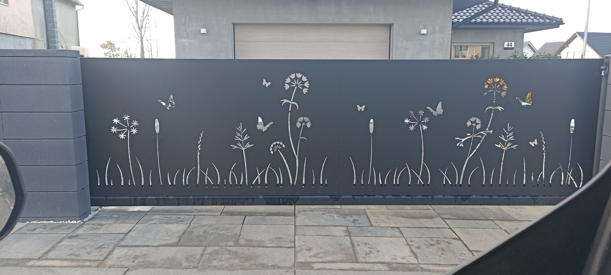 Nowoczesna, przesuwna brama wjazdowa w kolorze grafitowym z laserowo wycinanymi motywami roślinnymi i motylami, na tle kostki brukowej i domu z garażem.