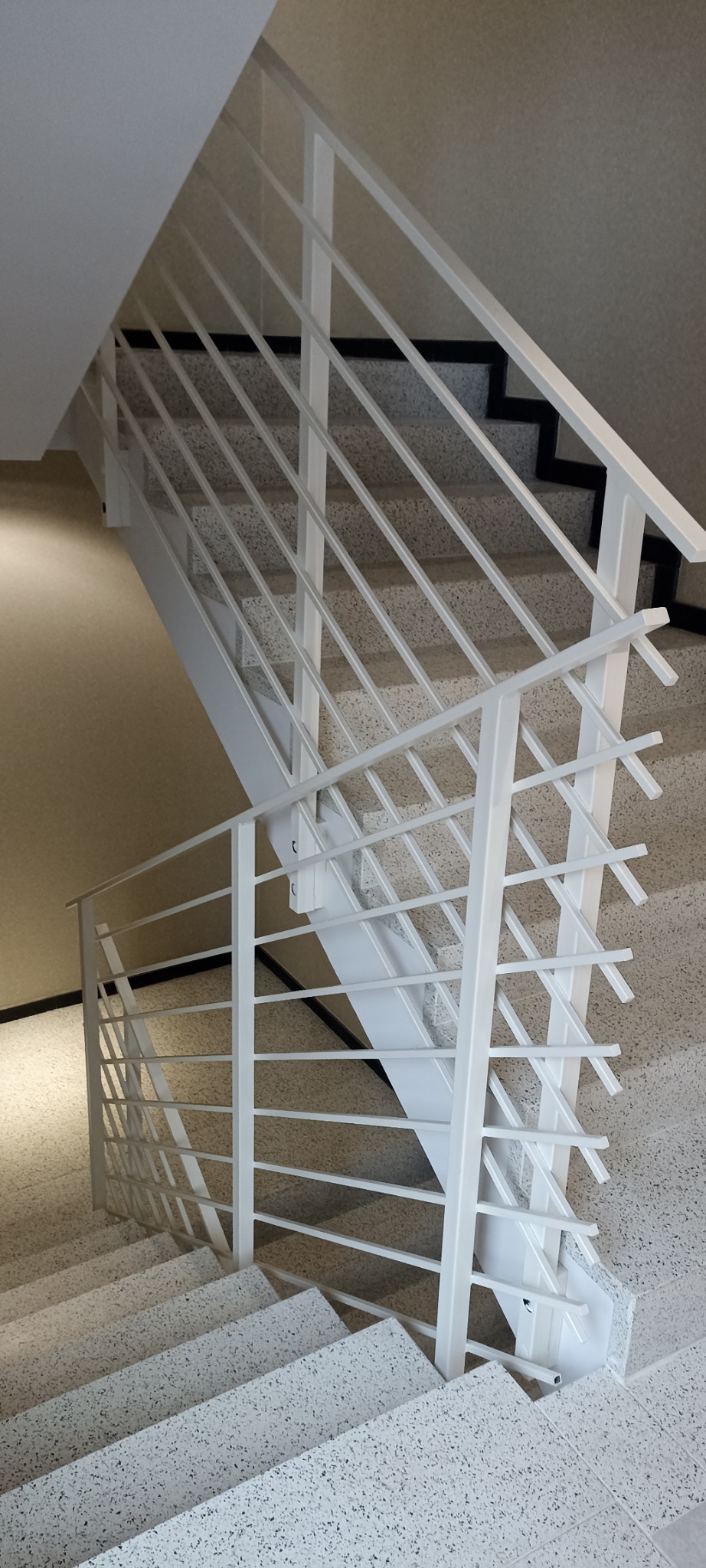 Białe, metalowe schody z minimalistyczną balustradą wewnątrz budynku, widok z góry na dół, schody wykończone lastryko.