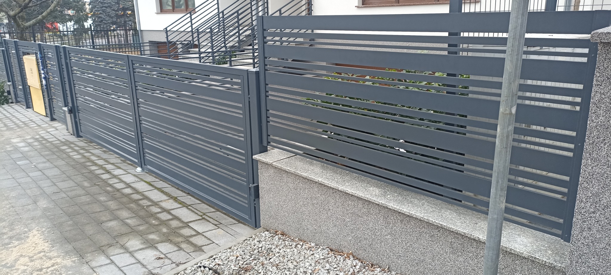 Nowoczesne, poziome, szare przęsła ogrodzeniowe z widocznymi elementami konstrukcyjnymi i słupkami z szarego kamienia, oddzielające posesję od chodnika z kostki brukowej.