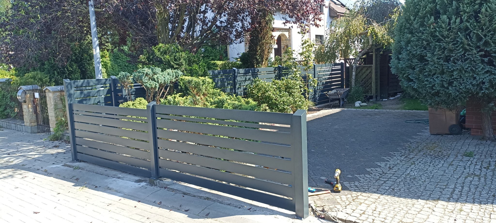 Nowoczesne, poziome ogrodzenie panelowe w kolorze szarym, z widocznymi narzędziami montażowymi na kostce brukowej, otoczone zielenią i zabudową jednorodzinną.