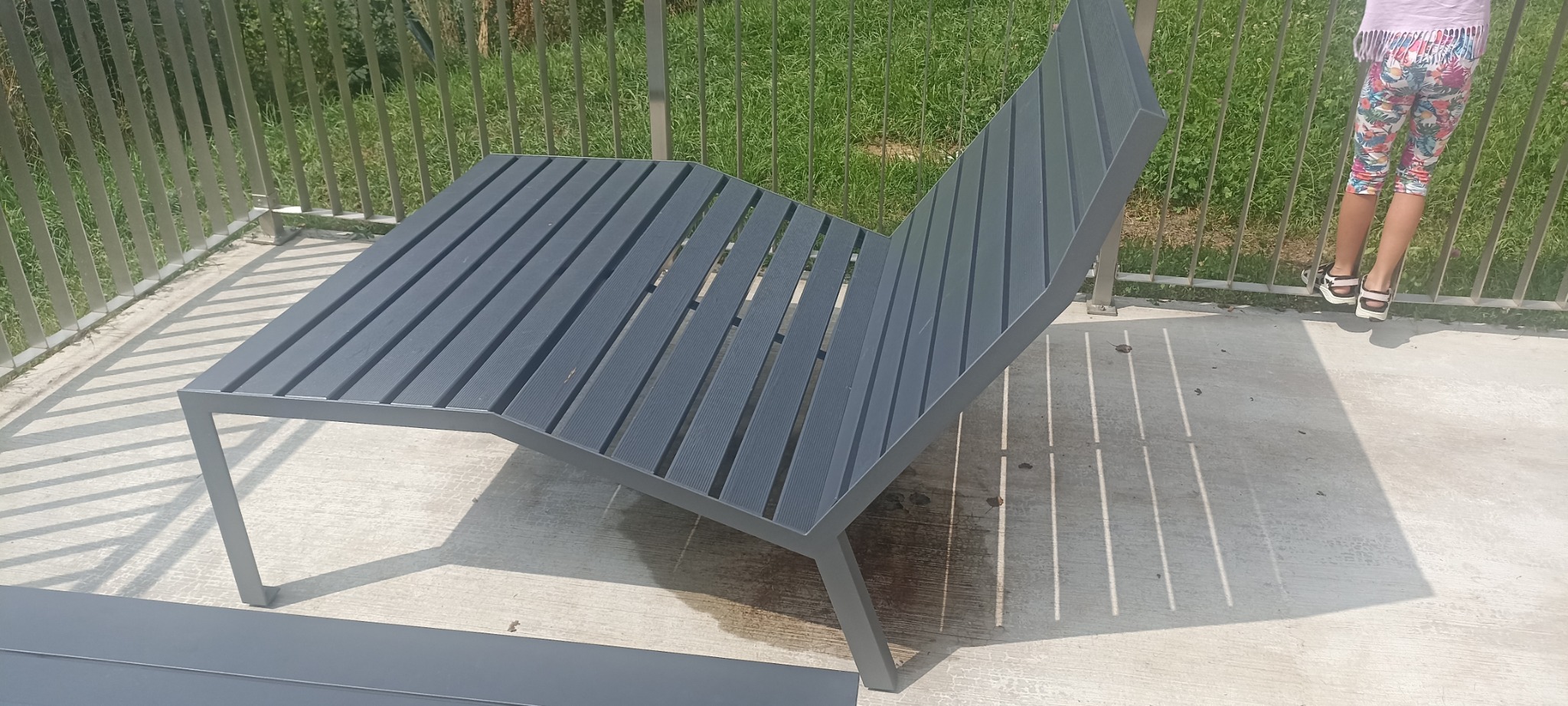 Nowoczesny, szary leżak ogrodowy z metalową ramą i drewnianymi listwami, rzucający cienie na betonowe podłoże, z widocznym fragmentem metalowej balustrady w tle i fragmentem osoby w kolorowych...