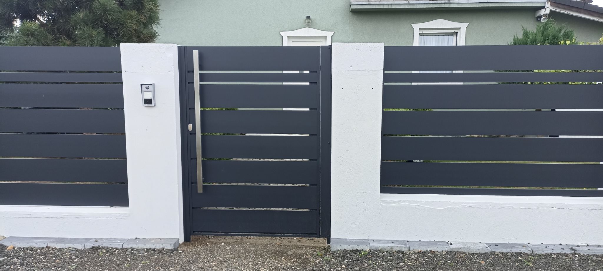Nowoczesna, antracytowa brama wjazdowa z poziomymi panelami, osadzona między białymi, murowanymi słupkami z domofonem, na tle zielonego domu.