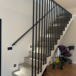 MARIUSZ GONDEK MAR-MONT - Schody wewnętrzne z szarymi stopniami i czarną, minimalistyczną balustradą metalową, oświetlenie schodowe w ścianie, na podłodze zabawki i wózek dziecięcy.