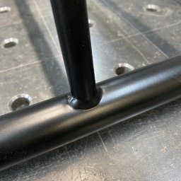 MARIUSZ GONDEK MAR-MONT - Spoina łącząca pionowy i poziomy element metalowej konstrukcji pomalowanej proszkowo na czarno, widoczna na tle stołu spawalniczego z otworami.