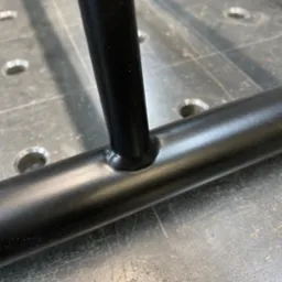 Spoina łącząca pionowy i poziomy element metalowej konstrukcji pomalowanej proszkowo na czarno, widoczna na tle stołu spawalniczego z otworami.