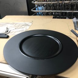 MARIUSZ GONDEK MAR-MONT - Czarny, okrągły element metalowy z wyraźną fakturą drewna, prawdopodobnie stopień schodowy, leży na kartonowym szablonie w warsztacie, w tle widoczne metalowe rury i konstrukcje.