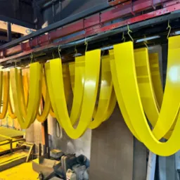 Żółte, metalowe elementy w kształcie podkowy zawieszone na wieszakach podczas procesu malowania proszkowego w hali produkcyjnej.