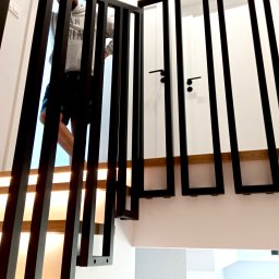 MARIUSZ GONDEK MAR-MONT - Nowoczesna, czarna balustrada schodowa z pionowych profili, zamontowana na drewnianych stopniach podświetlonych od spodu, osoba w tle wchodzi po schodach.