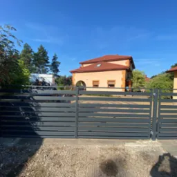 Nowoczesna, grafitowa brama przesuwna z poziomymi panelami, widok z zewnątrz na posesję z kostką brukową i domem z pomarańczową dachówką w tle.