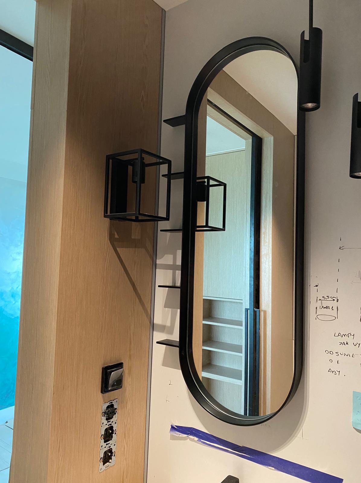 Nowoczesne oświetlenie wnętrza: czarne, geometryczne kinkiety i podłużne lustro z półkami na ścianie z instalacją elektryczną w trakcie montażu.