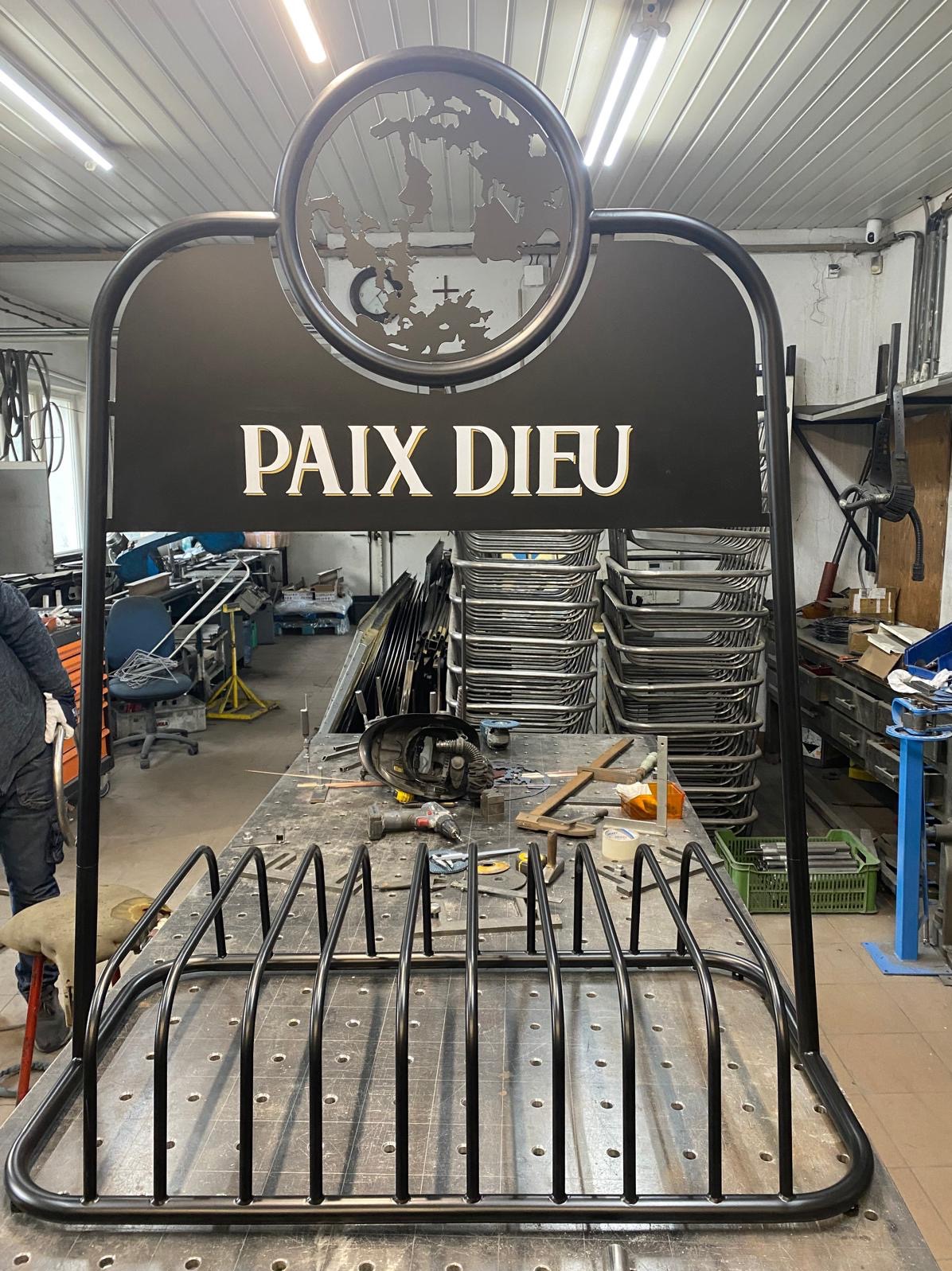 Metalowy stojak na rowery z napisem 'PAIX DIEU' i wyciętą mapą świata w okręgu, widoczny w warsztacie z narzędziami i stosem metalowych konstrukcji w tle.