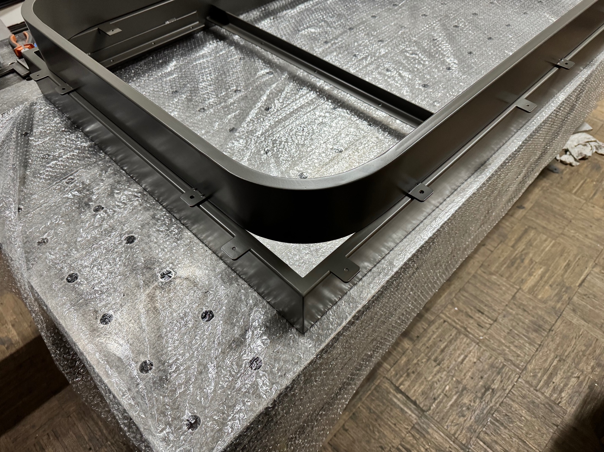 Metalowa rama z zaokrąglonym rogiem, pomalowana na szaro, leżąca na folii bąbelkowej, widoczne mocowania.