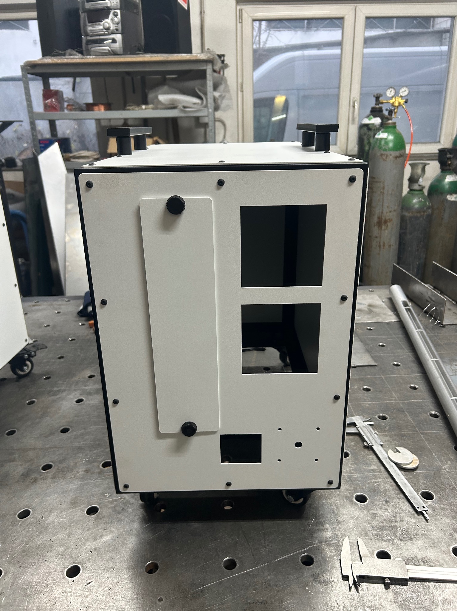 Metalowa obudowa urządzenia na kółkach z otworami, umieszczona na stole spawalniczym, obok narzędzi pomiarowych i butli z gazem.