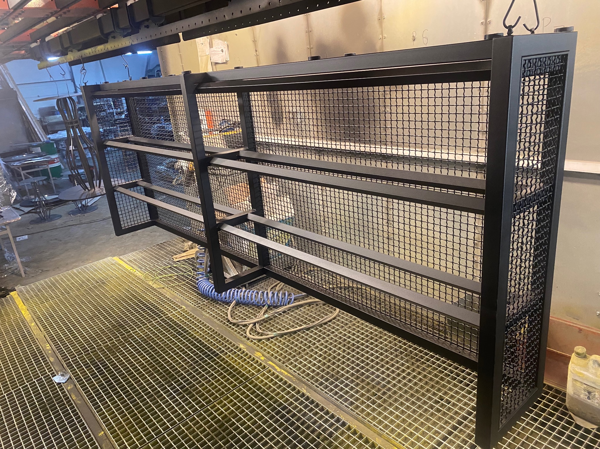 Metalowa konstrukcja z siatki, czarna, wisząca na haku w hali produkcyjnej, z widocznym przewodem pneumatycznym i kratą podłogową.