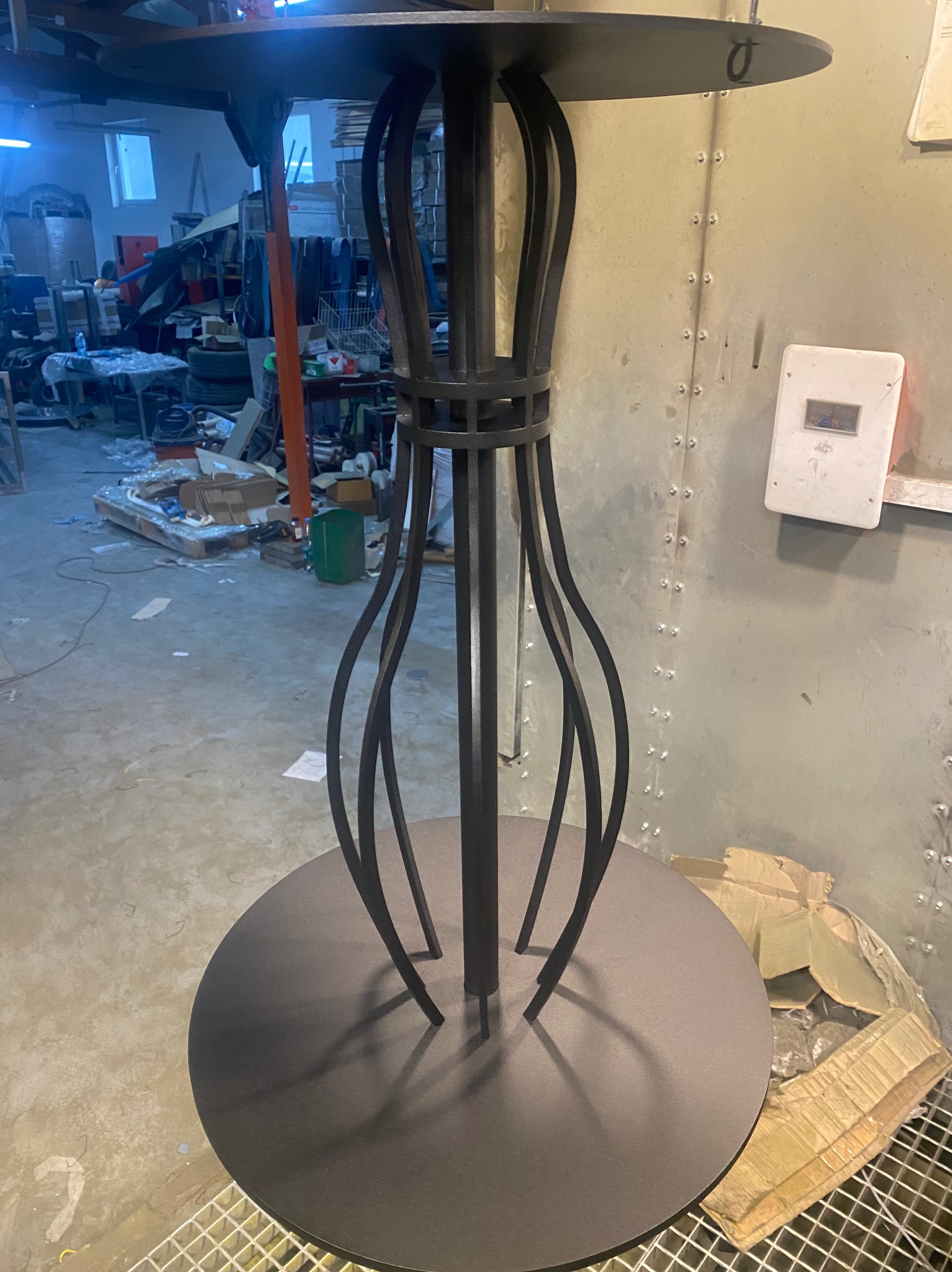 Stół barowy z metalową konstrukcją w kształcie klepsydry, wykonany z czarnej stali, widoczny w warsztacie z rozrzuconymi materiałami i narzędziami w tle.