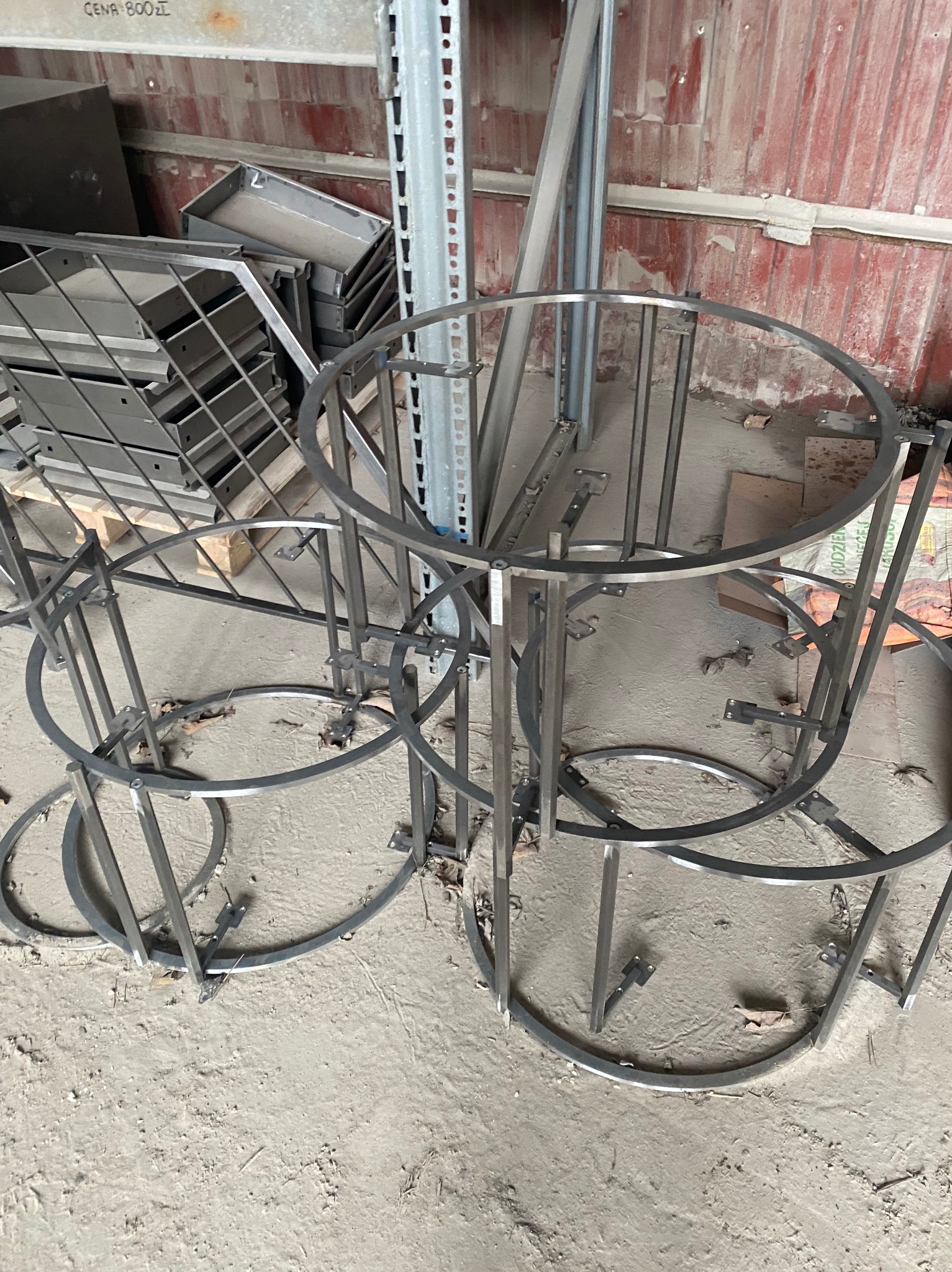 Stalowe, surowe konstrukcje z okrągłych profili, prawdopodobnie elementy balustrady lub schodów, leżące na betonowej podłodze w warsztacie, w tle blaszana ściana i paleta z metalowymi elementami.