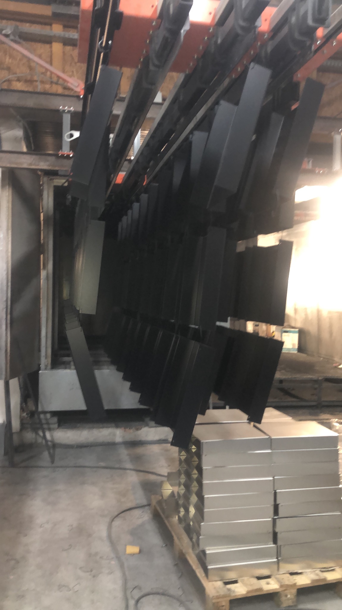 Elementy metalowe zawieszone na linii produkcyjnej, prawdopodobnie w trakcie malowania proszkowego, z paletą gotowych, niepomalowanych elementów w tle.