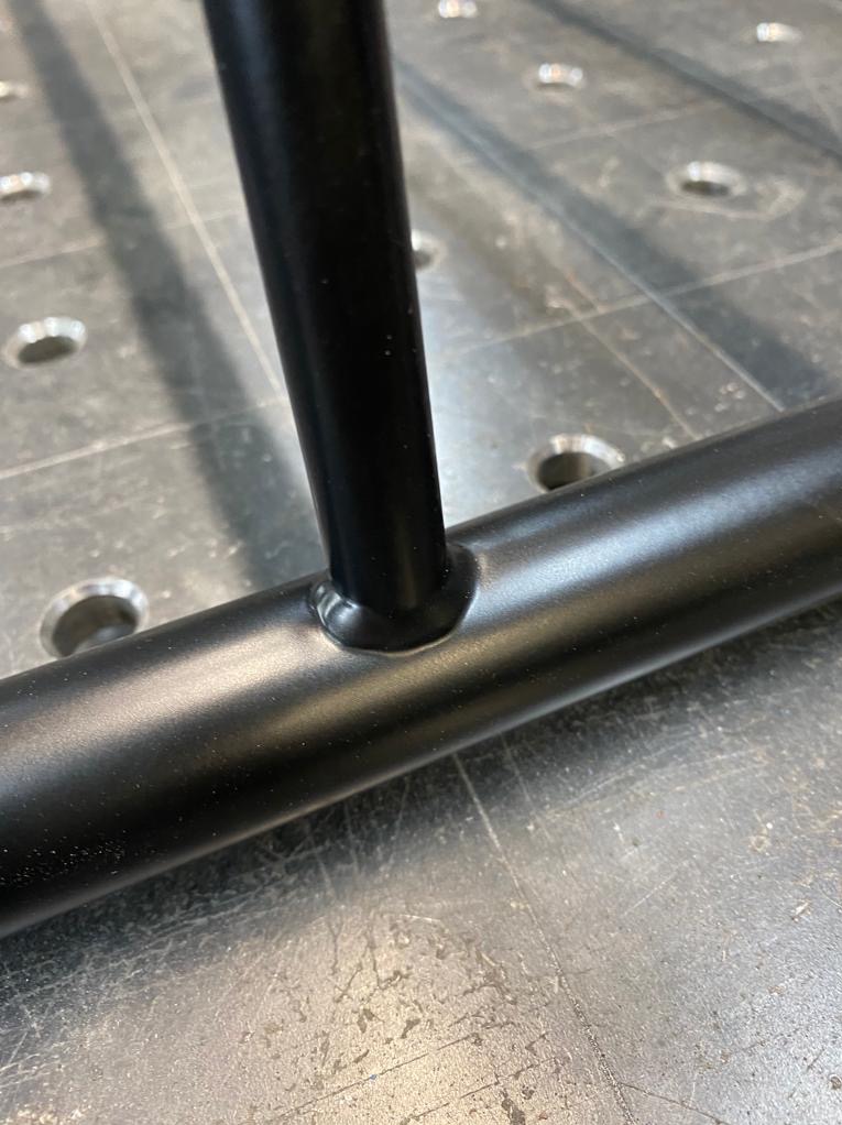 Spoina łącząca pionowy i poziomy element metalowej konstrukcji pomalowanej proszkowo na czarno, widoczna na tle stołu spawalniczego z otworami.