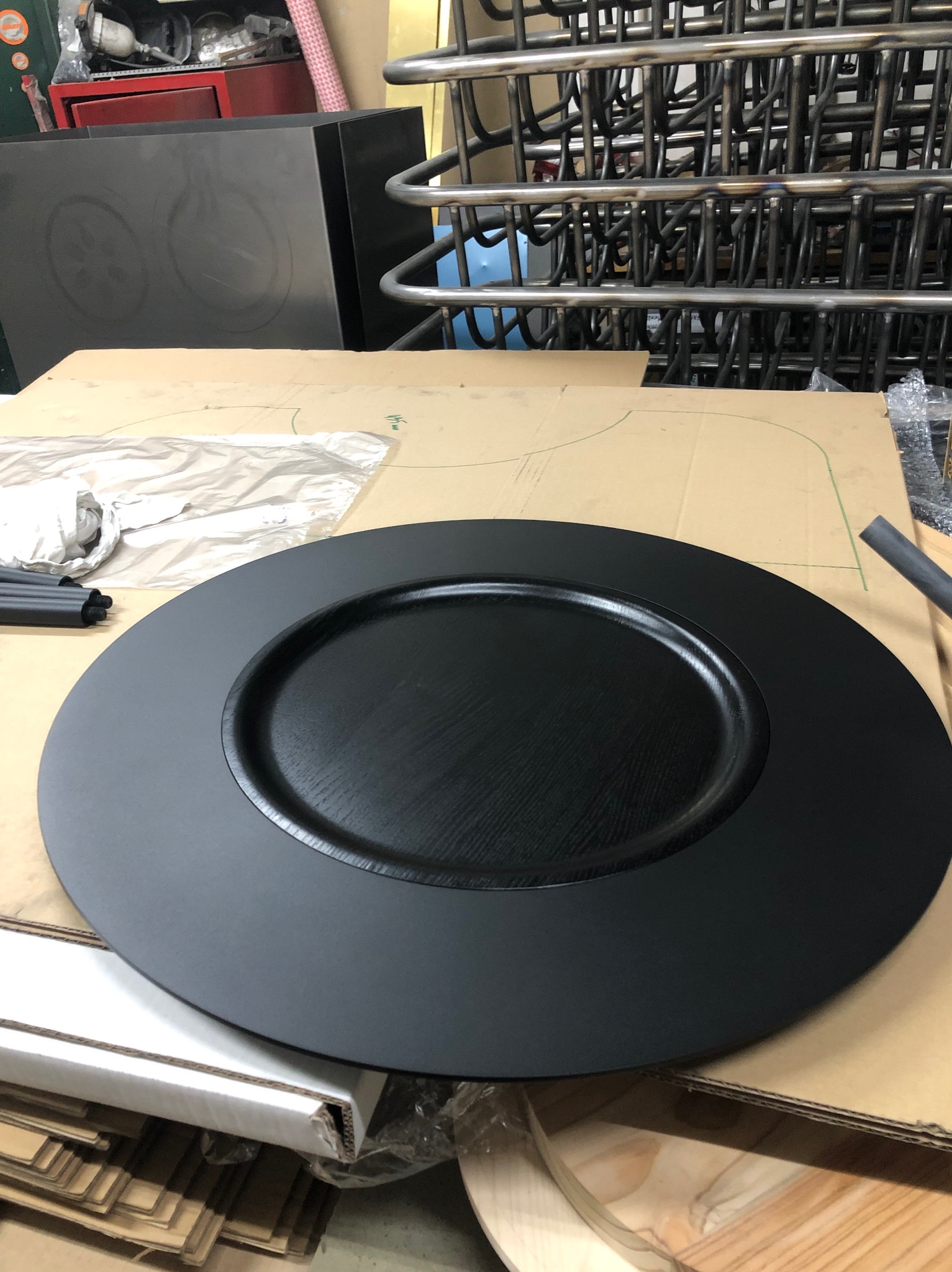 Czarny, okrągły element metalowy z wyraźną fakturą drewna, prawdopodobnie stopień schodowy, leży na kartonowym szablonie w warsztacie, w tle widoczne metalowe rury i konstrukcje.