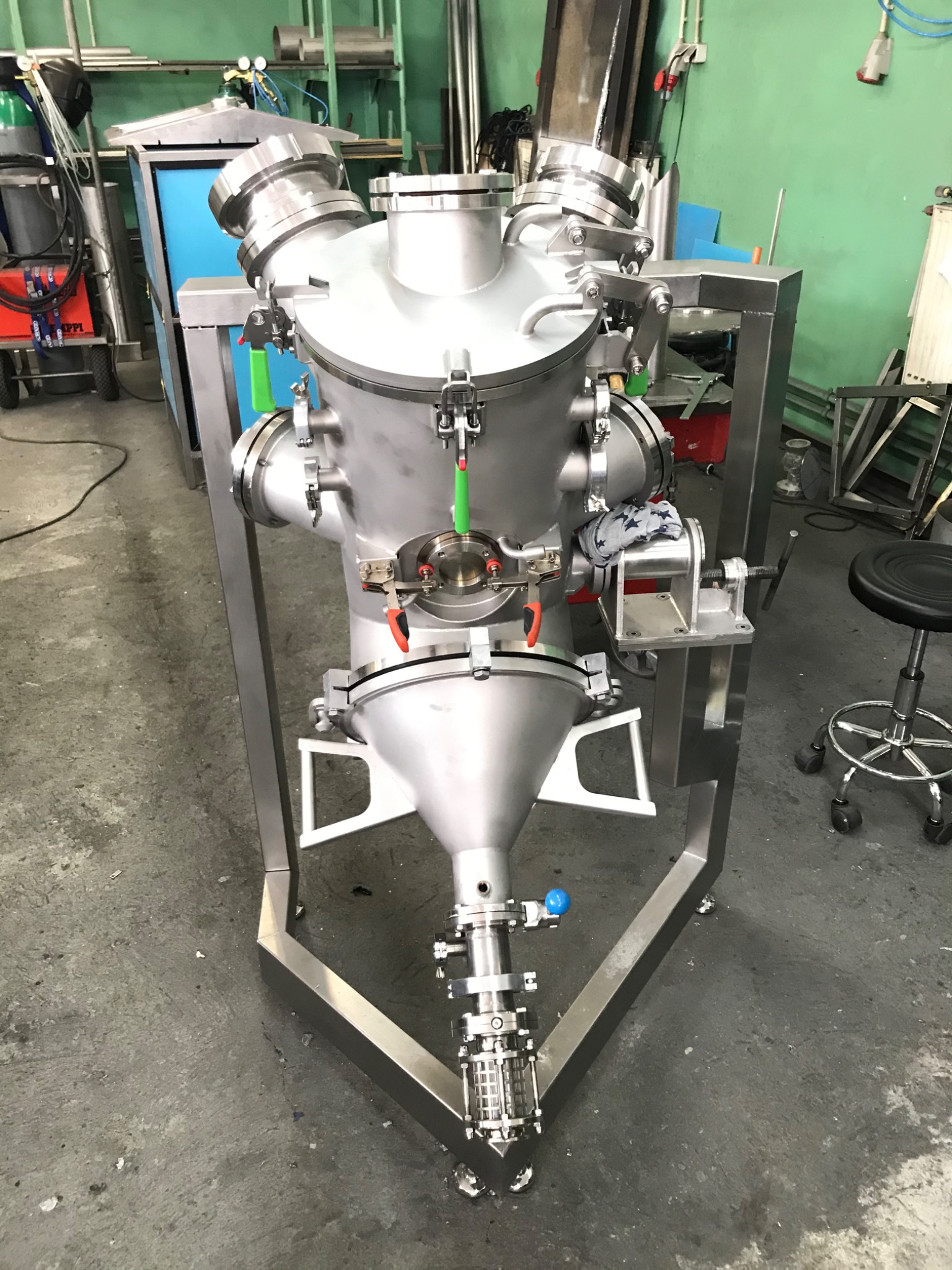 Srebrna, metalowa konstrukcja przemysłowa z licznymi złączami i zaworami, umieszczona na trójkątnej podstawie w warsztacie.