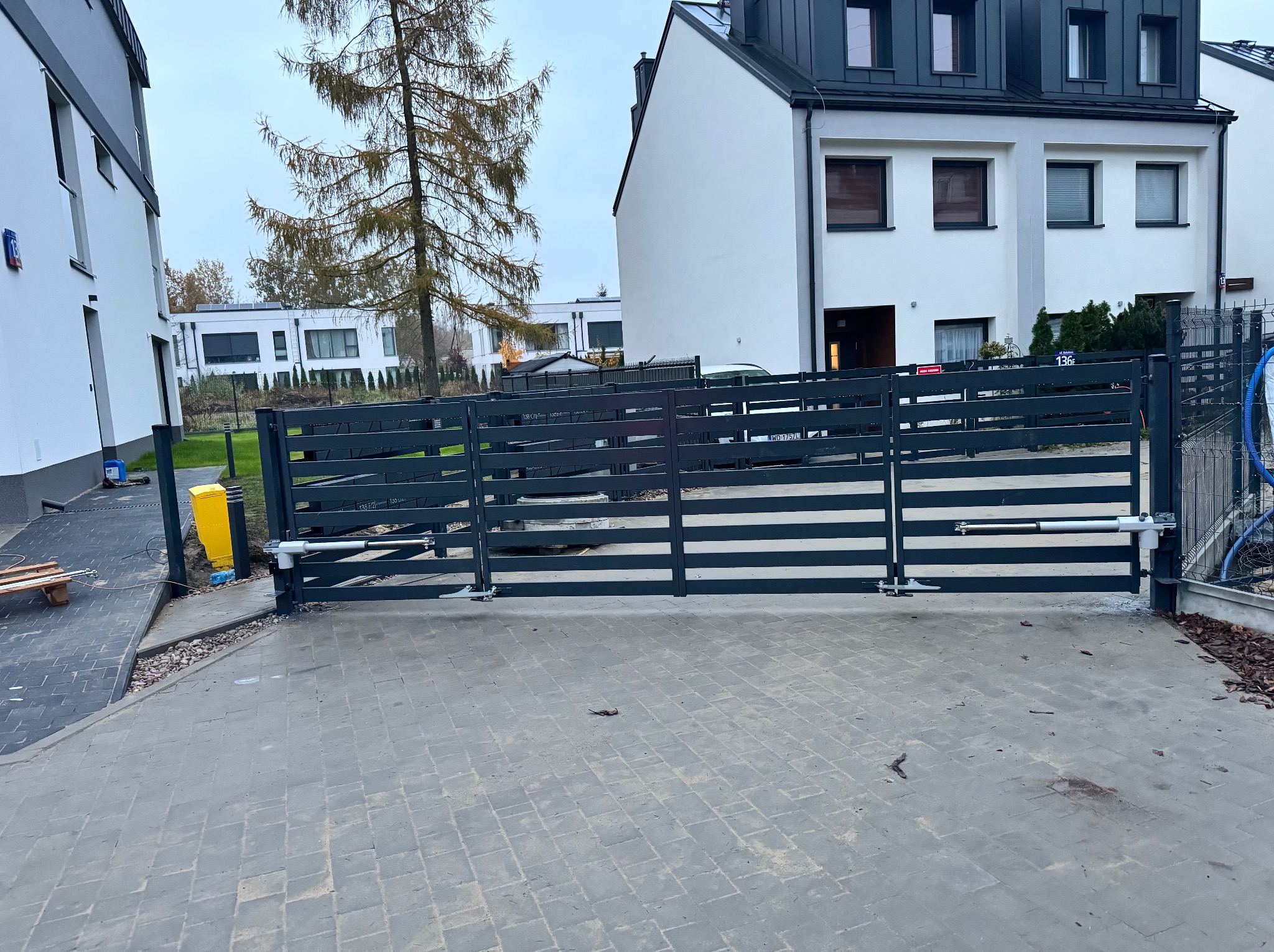 Nowoczesna, pozioma brama wjazdowa z automatycznym napędem, zamontowana przed szeregową zabudową mieszkalną w szarej kolorystyce z kostki brukowej.