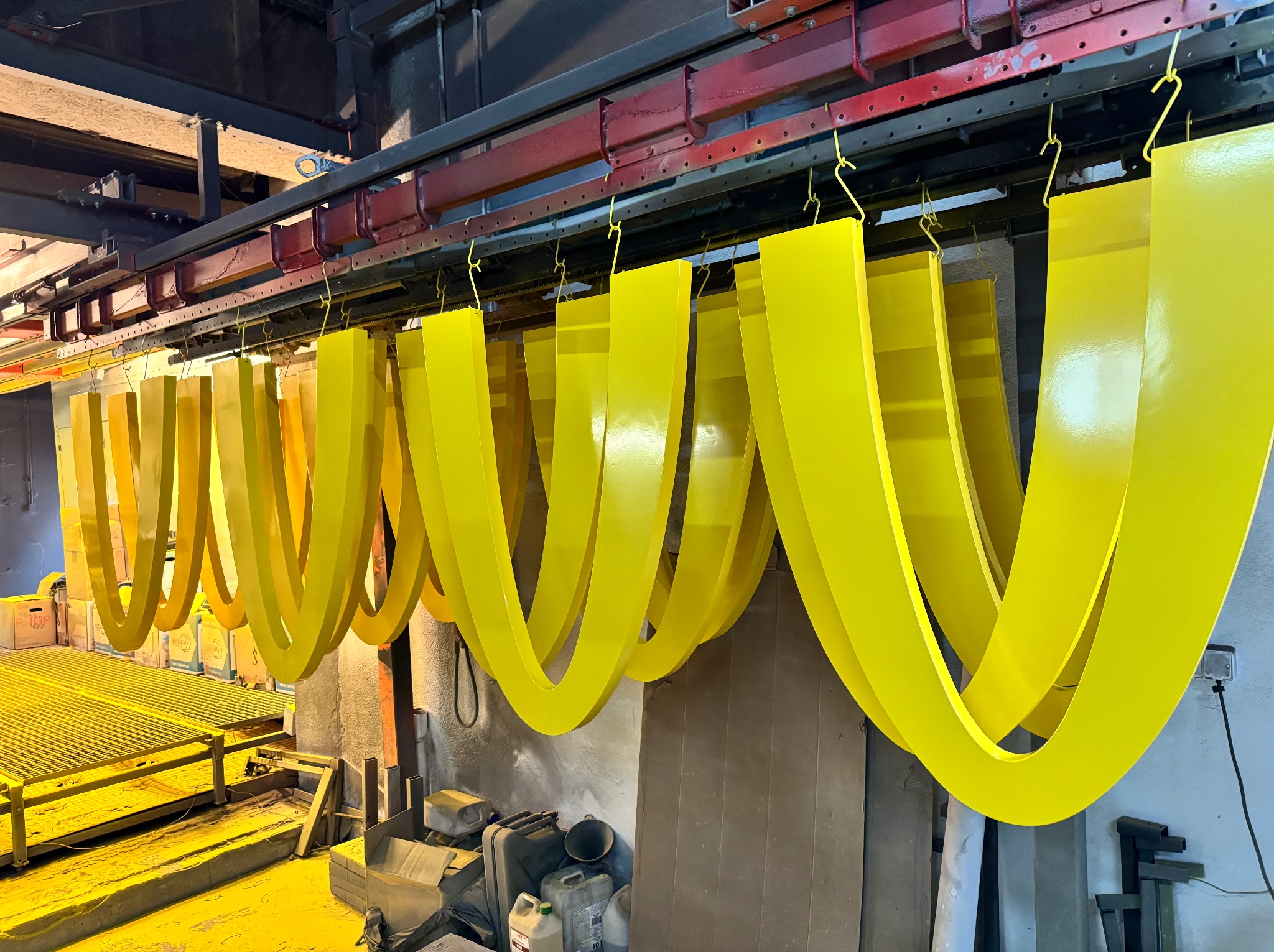 Żółte, metalowe elementy w kształcie podkowy zawieszone na wieszakach podczas procesu malowania proszkowego w hali produkcyjnej.