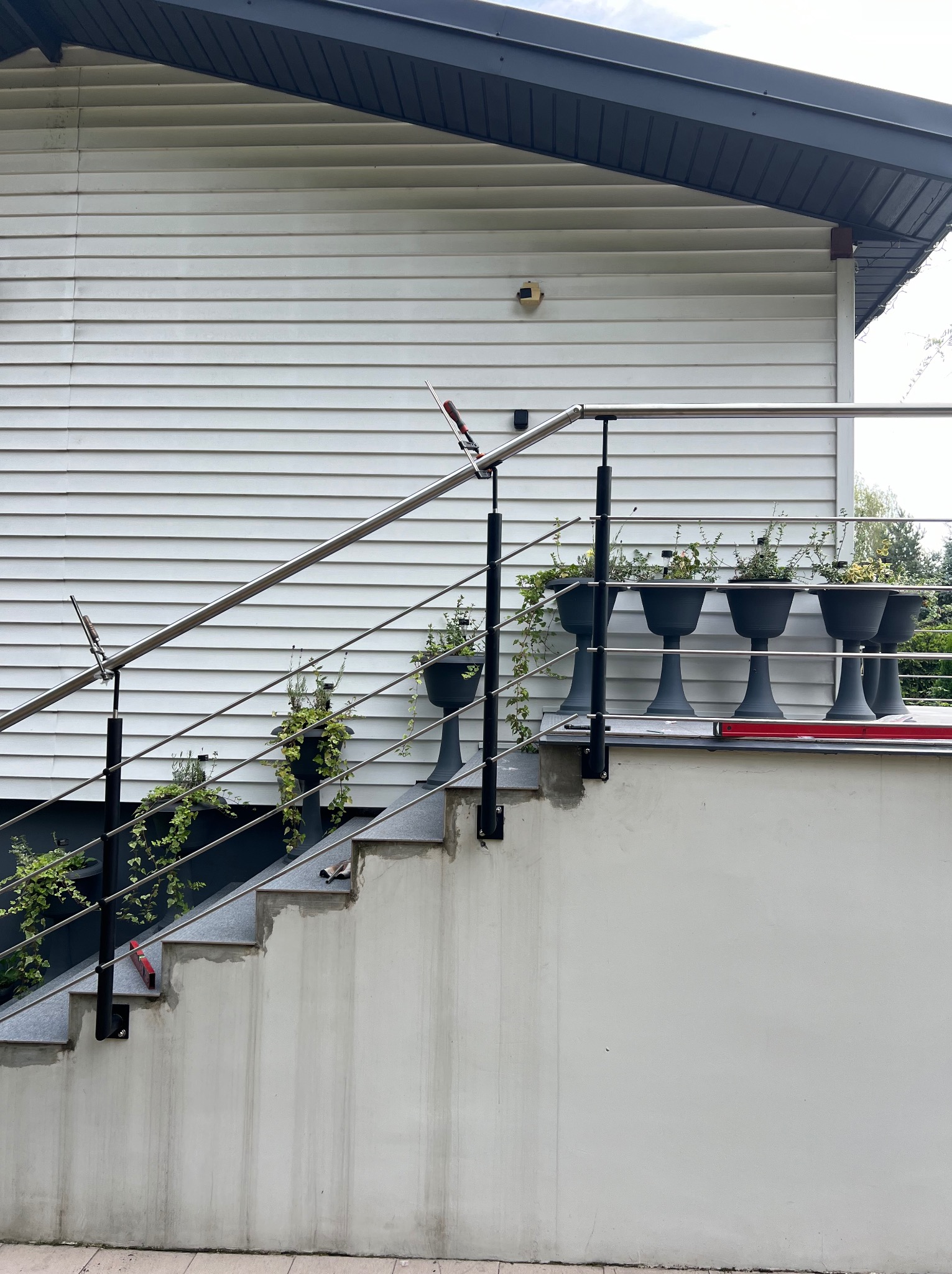 Zewnętrzne schody z szarymi stopniami i metalową balustradą z nierdzewnej stali, na której zamontowano czarne donice z roślinami, widoczne ślady prac wykończeniowych na betonie.
