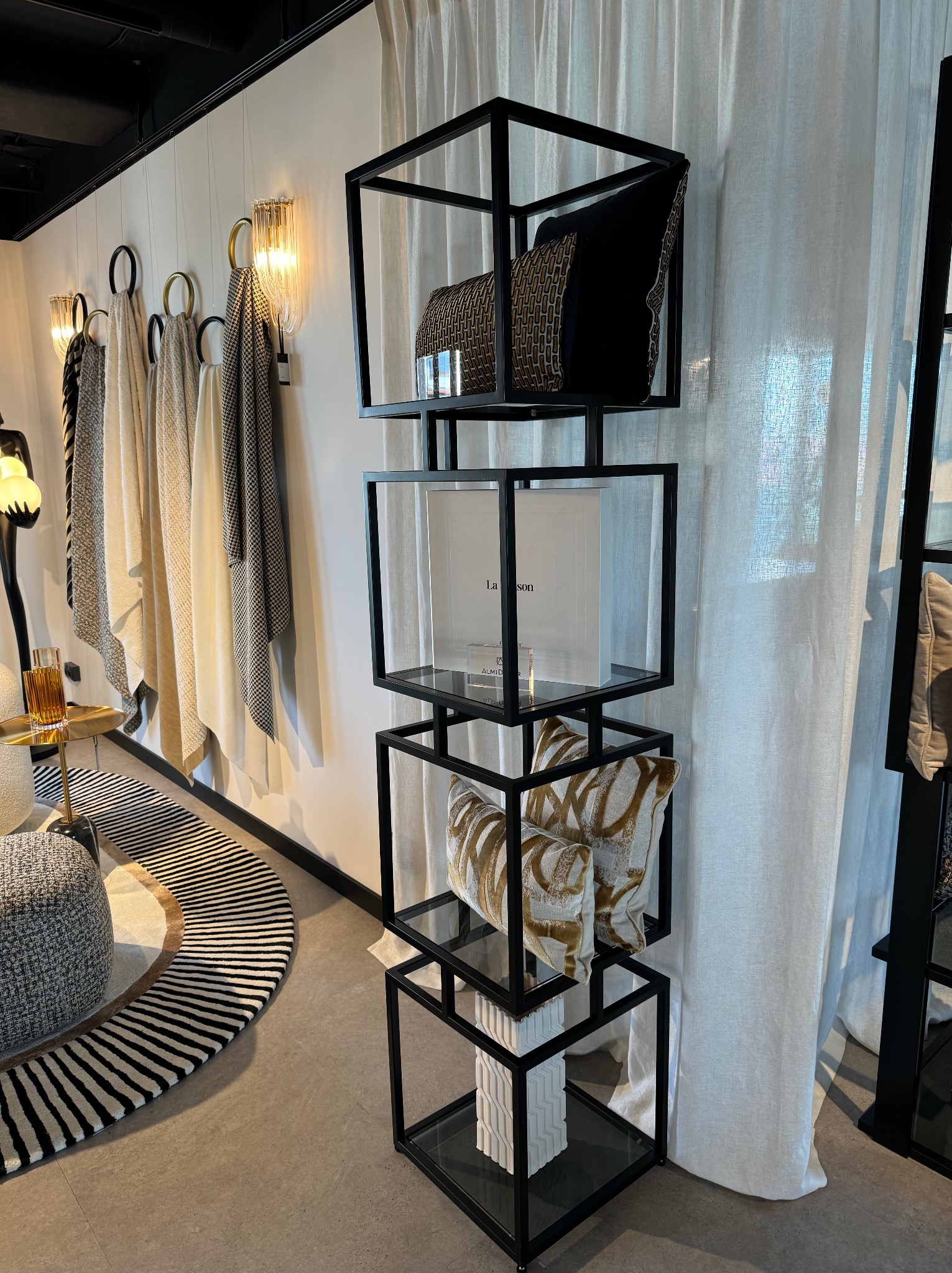 Nowoczesny regał metalowy z półkami szklanymi, prezentujący dekoracyjne poduszki i książkę w minimalistycznym wnętrzu z zasłonami i próbkami tkanin na ścianie.