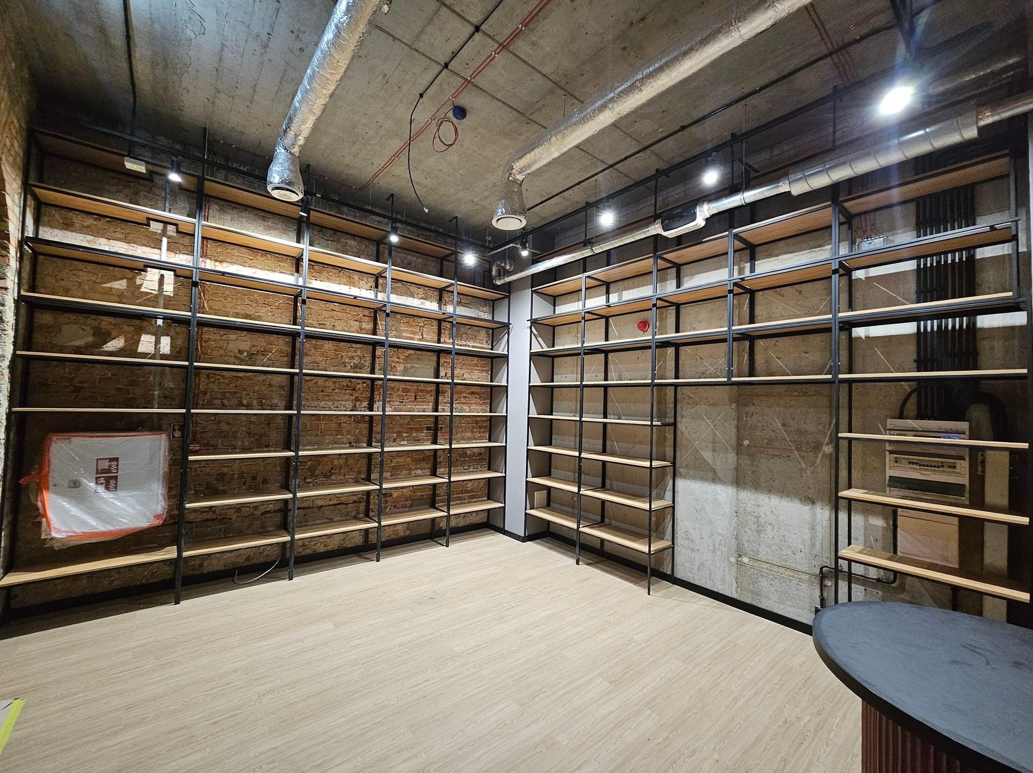 Metalowa konstrukcja regałów z drewnianymi półkami, wykonana w Nadarzynie, na tle surowej cegły i betonu, widoczne instalacje pod sufitem.