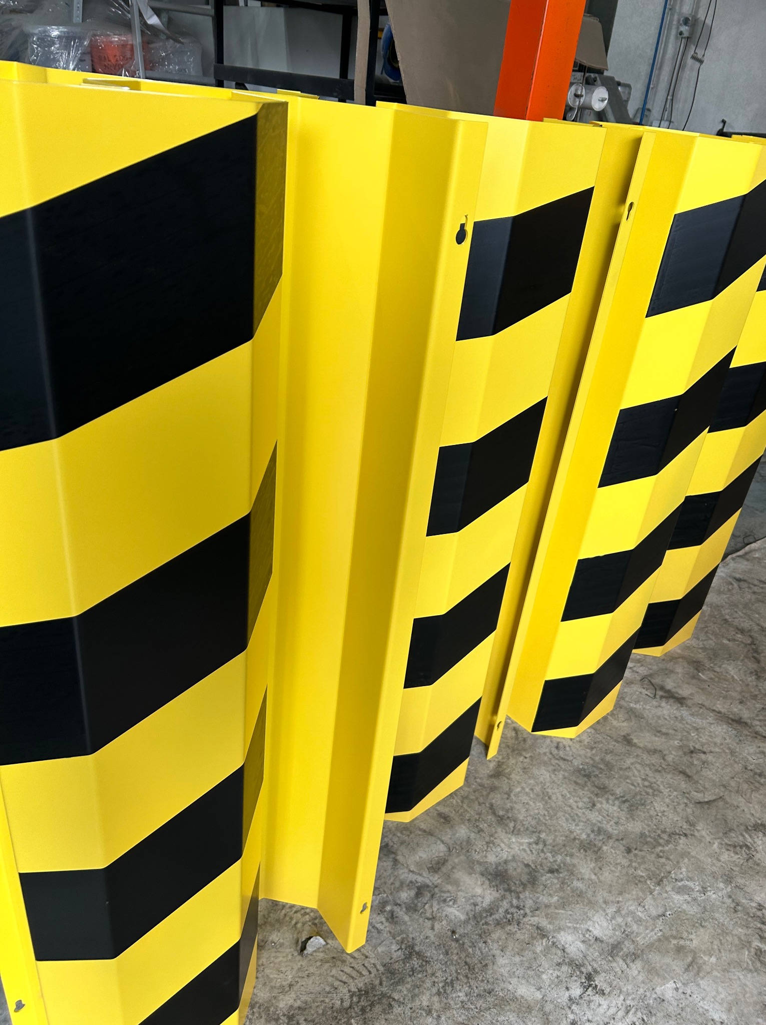Żółto-czarne, metalowe odbojniki magazynowe, ustawione jeden za drugim, z widocznymi otworami montażowymi, na szarym, betonowym podłożu.