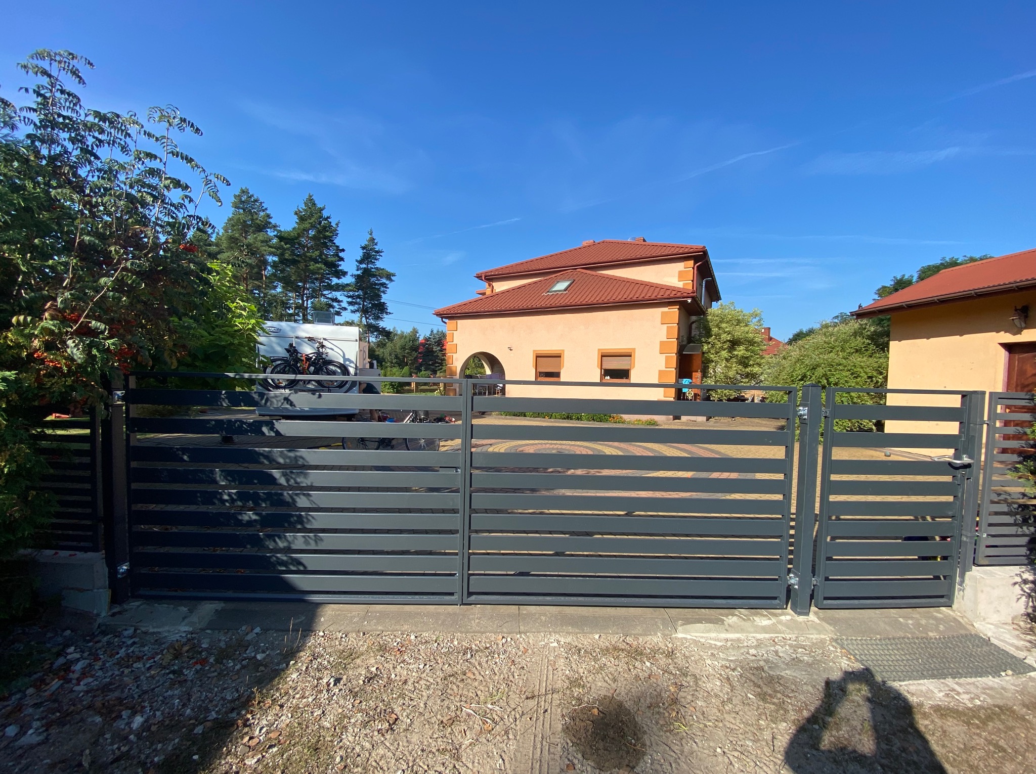 Nowoczesna, grafitowa brama przesuwna z poziomymi panelami, widok z zewnątrz na posesję z kostką brukową i domem z pomarańczową dachówką w tle.