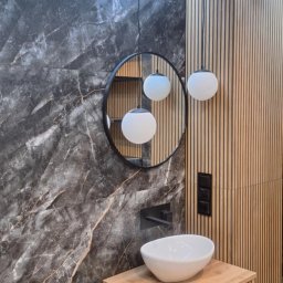 Zomar - Nowoczesna łazienka z marmurową ścianą, okrągłym lustrem i drewnianą szafką pod umywalką. Wiszące lampy kuliste dodają elegancji. Widoczne detale wykończenia.