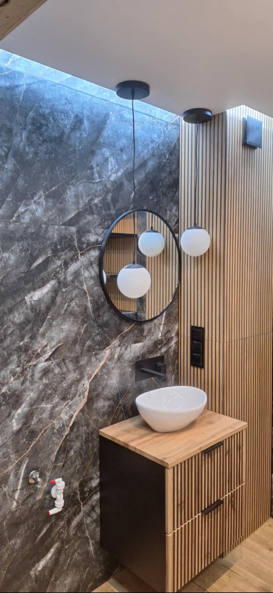 Nowoczesna łazienka z marmurową ścianą, okrągłym lustrem i drewnianą szafką pod umywalką. Wiszące lampy kuliste dodają elegancji. Widoczne detale wykończenia.
