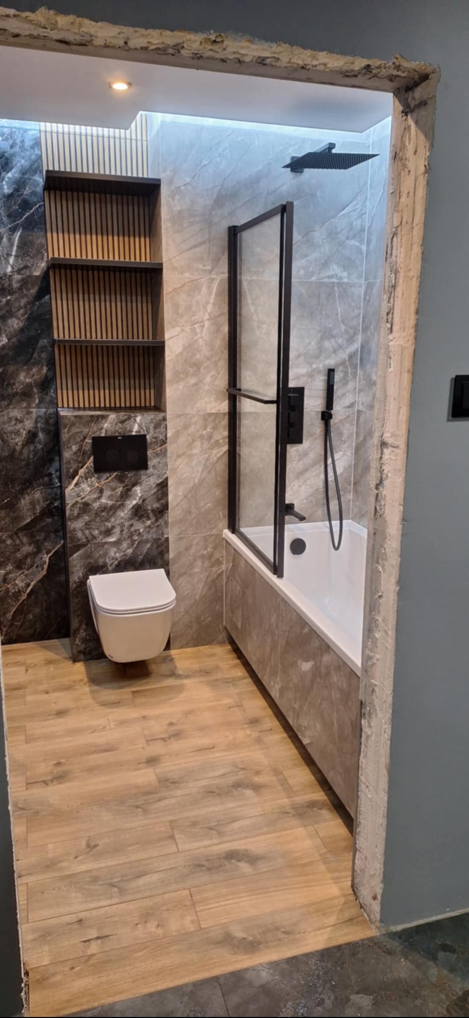 Nowoczesna łazienka po remoncie: marmurowe płytki, drewnopodobna podłoga, czarna armatura i minimalistyczne półki. Widok zza futryny.