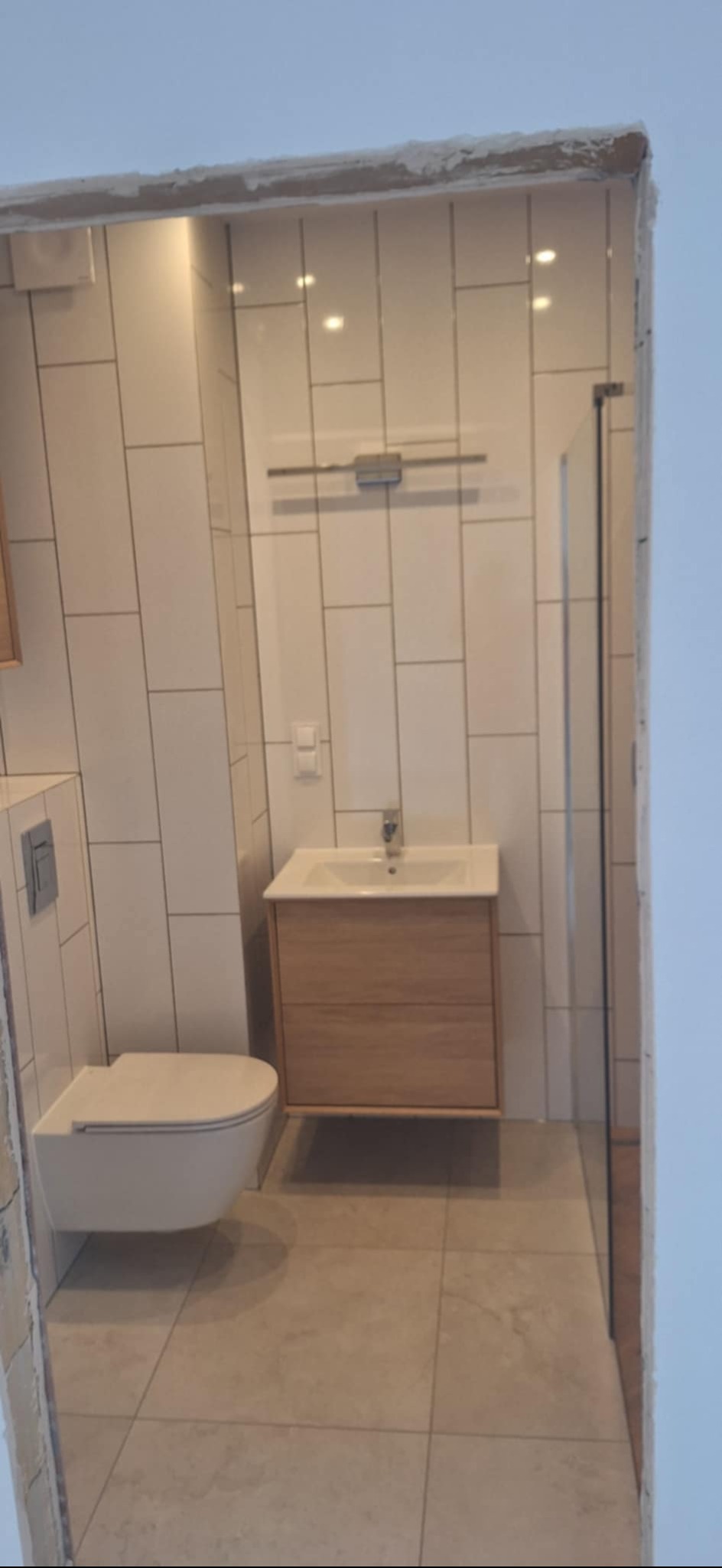 Nowoczesna łazienka z białymi, pionowo ułożonymi płytkami, drewnianą szafką pod umywalką i podwieszaną toaletą. Minimalistyczny design, jasne oświetlenie.