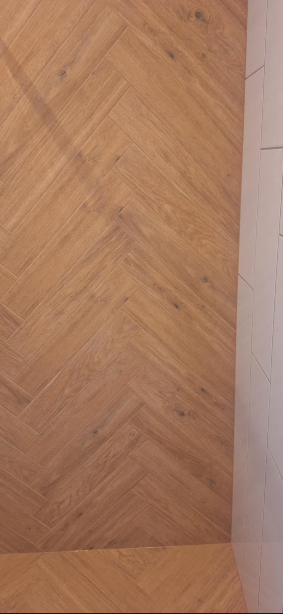 Ułożona parkietowa podłoga z drewnopodobnych paneli w jodełkę, sąsiadująca z białą, pionowo ułożoną glazurą ścienną. Widok z góry, ciepłe oświetlenie.