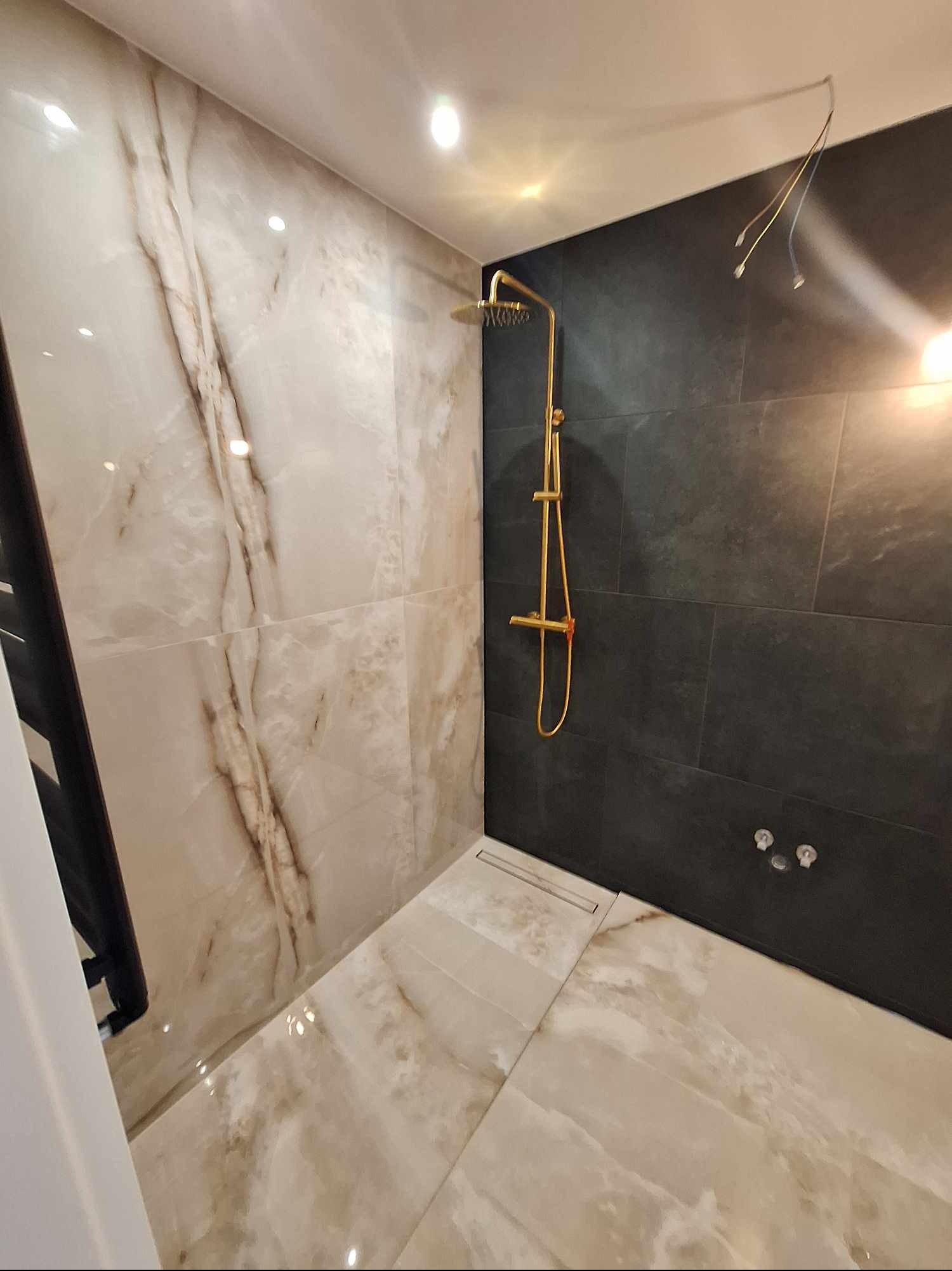 Nowoczesna łazienka po remoncie: beżowe płytki imitujące marmur, czarna ściana z złotą armaturą prysznicową. Widoczne niedokończone podłączenie oświetlenia.