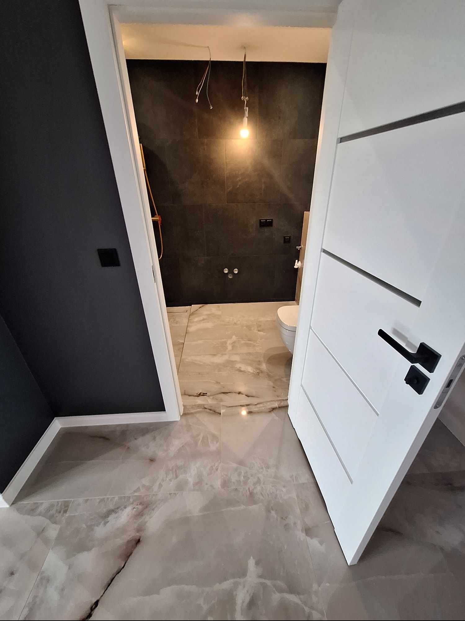 Nowoczesna łazienka z szarą glazurą, marmurową podłogą i białą, częściowo otwartą, designerską  drzwią z czarną klamką. Widok z zewnątrz, oświetlenie górne.