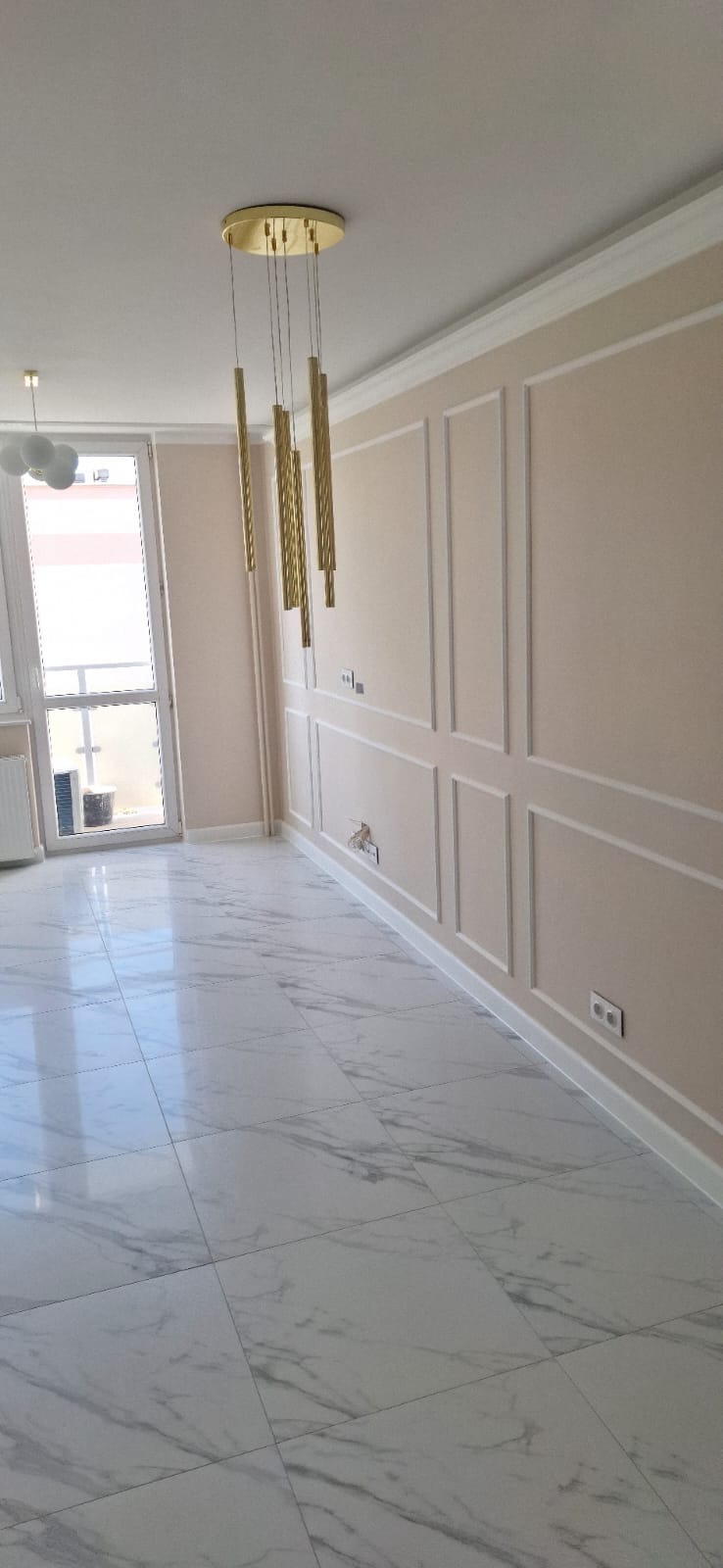 Wykończone wnętrze: marmurowe płytki podłogowe, ściany w odcieniach beżu z dekoracyjnymi listwami, złota lampa wisząca. Widoczne gniazdka elektryczne i okno balkonowe.