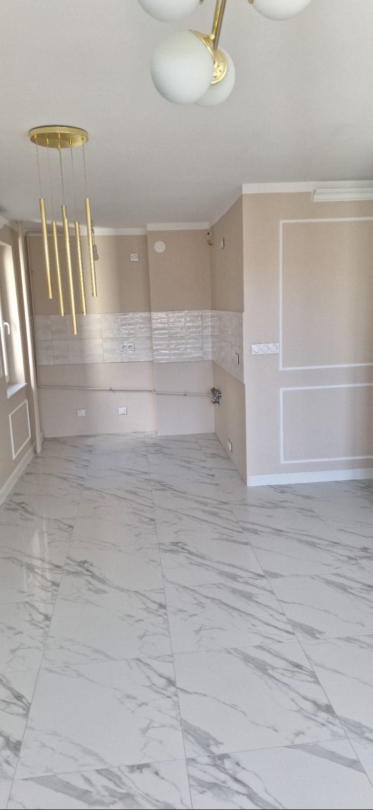 Jasne wnętrze po remoncie: białe płytki imitujące marmur na podłodze, beżowe ściany z dekoracyjnymi listwami i fragmentem glazury nad blatem. Nowoczesne oświetlenie.