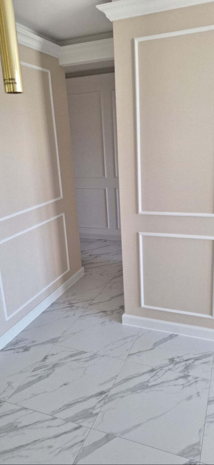 Eleganckie wykończenie wnętrza: beżowe ściany z białymi listwami dekoracyjnymi, marmurowe płytki podłogowe. Widok na kolejne pomieszczenie z podobnym stylem.