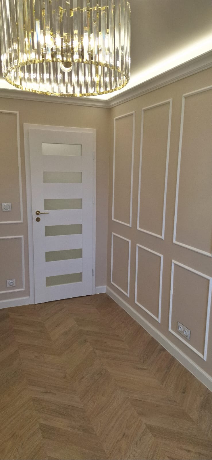 Eleganckie wnętrze z parkietem w jodełkę, białą stolarką drzwiową i dekoracyjnymi panelami ściennymi. Kryształowy żyrandol dodaje luksusowego charakteru.