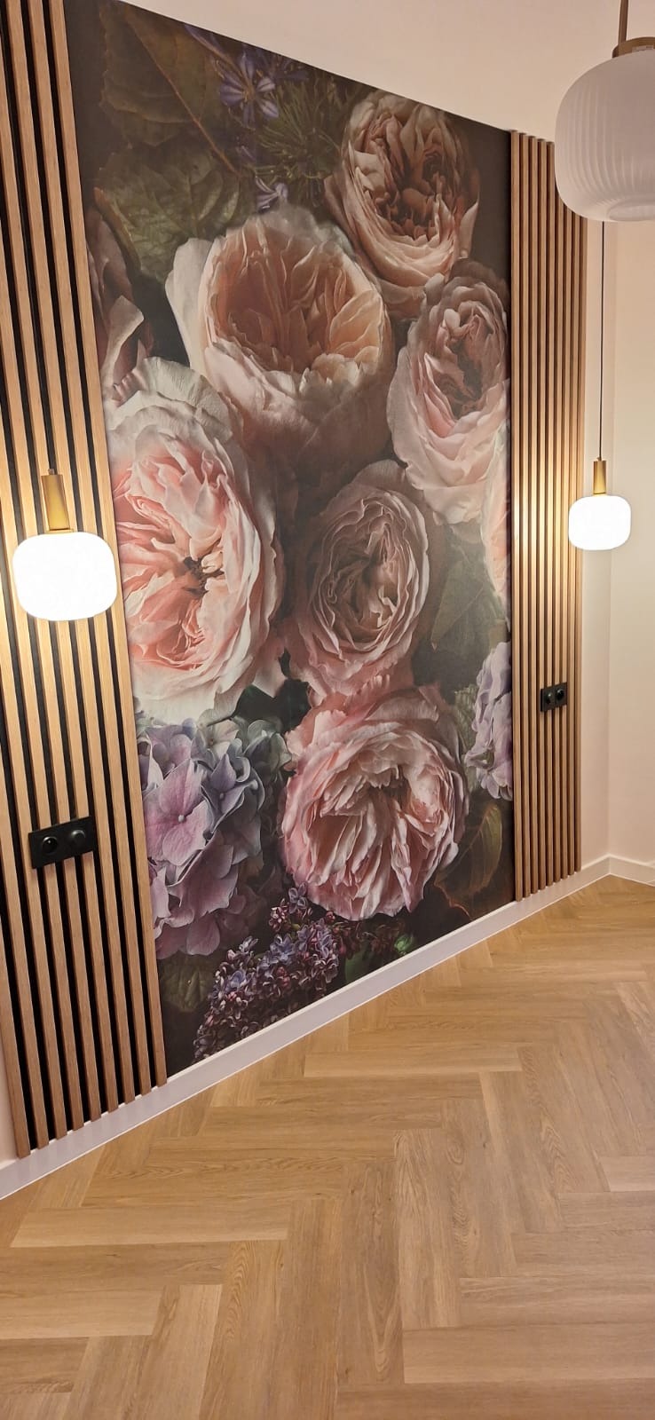 Wykończone wnętrze z fototapetą w kwiaty, drewnianymi lamelami na ścianie i parkietem ułożonym w jodełkę. Dwie lampy wiszące z białymi kloszami.