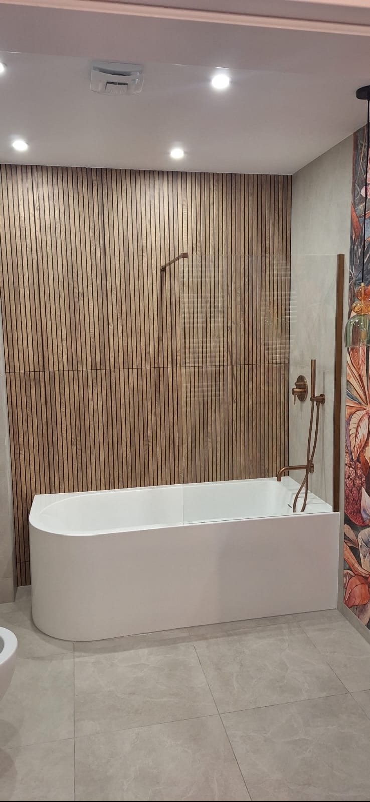 Nowoczesna łazienka z wanną, drewnianą ścianą z lameli i szklanym parawanem. Armatura w kolorze miedzi. Minimalistyczny design z akcentem roślinnym.