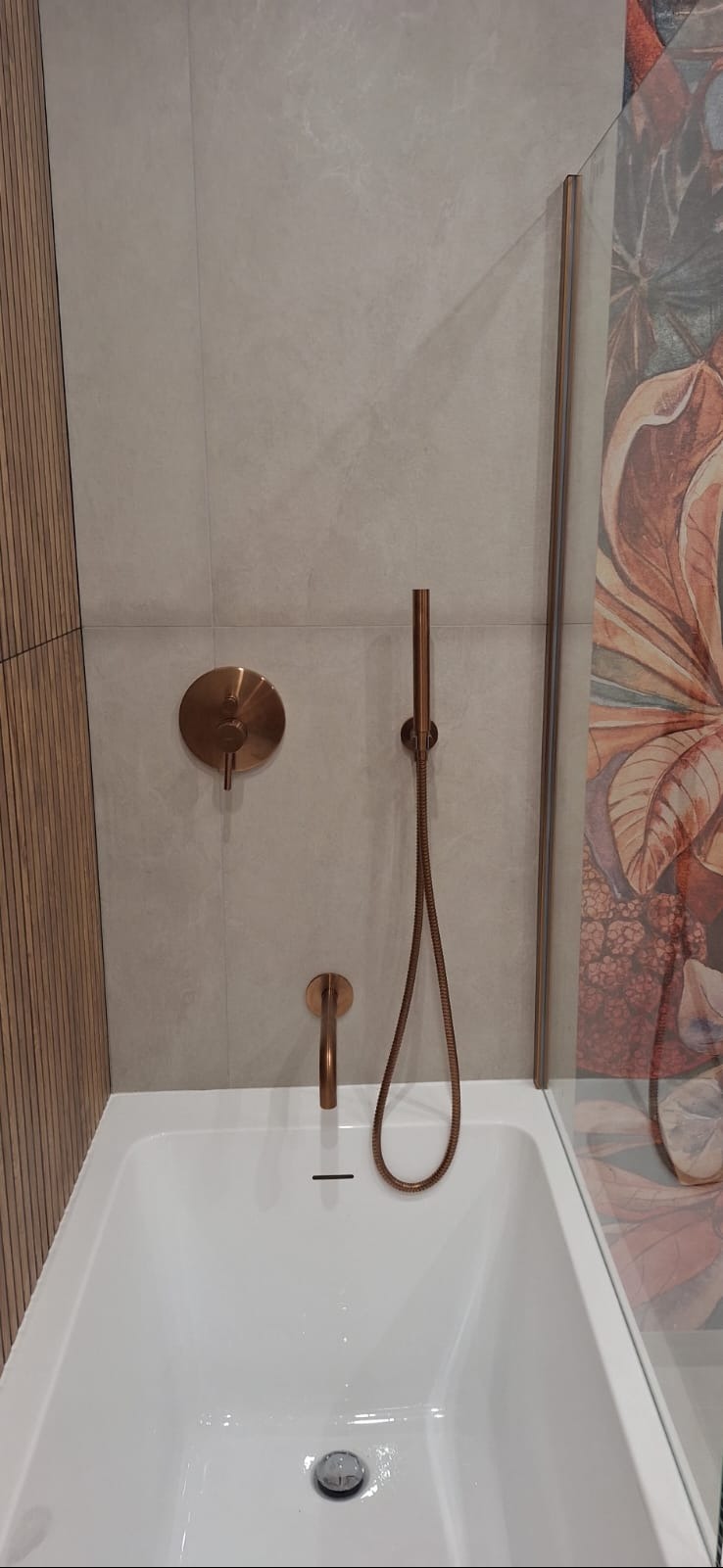 Nowoczesna łazienka z białą wanną, miedzianą armaturą i szklanym panelem z motywem roślinnym. Ściany wyłożone jasnymi płytkami, a obok wanny drewniane panele ścienne.