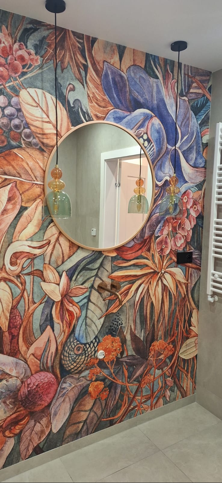 Wzorzysta tapeta w kwiaty i okrągłe lustro w łazience w Dzierżeninie. Wiszące lampy z kolorowego szkła. Grzejnik drabinkowy po prawej stronie.