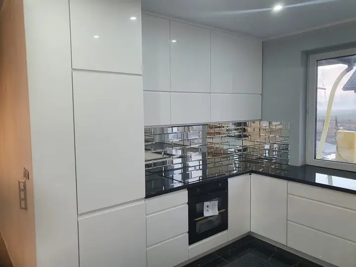 Biała, nowoczesna kuchnia z błyszczącymi frontami, czarnym blatem i lustrzanymi płytkami ułożonymi w cegiełkę nad piekarnikiem.