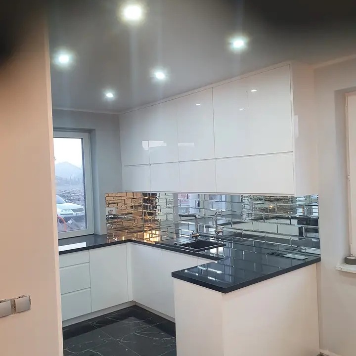 Biała kuchnia na wymiar z czarnym blatem i lustrzanymi płytkami na ścianie, widok na zewnątrz przez okno.