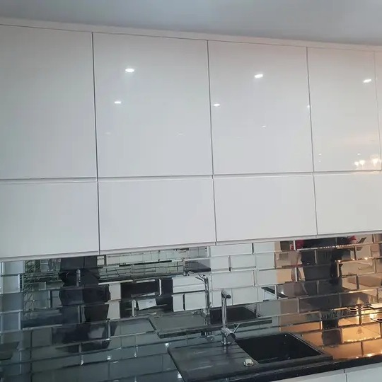 Białe, lakierowane szafki kuchenne wiszące nad blatem z czarnym zlewem i lustrzanymi płytkami w stylu cegiełek na ścianie.